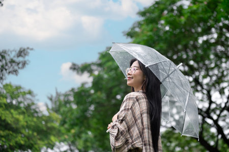 Young asian glasses woman holding transparent umbrella under the rain while standing in park or...