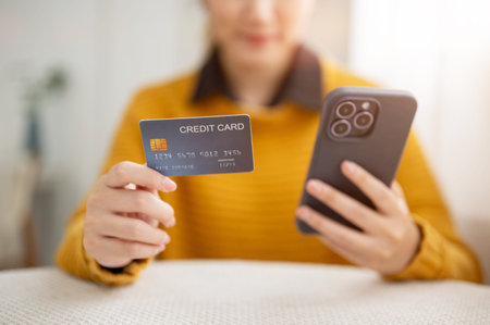 Close up of woman's hands holding smartphone and showing a credit card while sitting at table in...