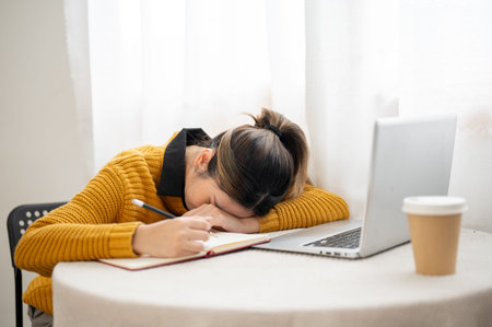 Asian woman student or a writer holding pencil over notebook sleeping aside laptop and coffee on...