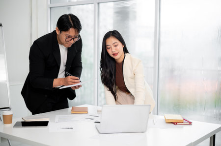 Businesswoman or office worker showing data on laptop for a man to note down over table with...