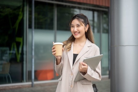 Beautiful asian woman holding a coffee cup and carrying a laptop while standing in front of the store. Working or Study, Urban life, Chilling Outside.の写真素材