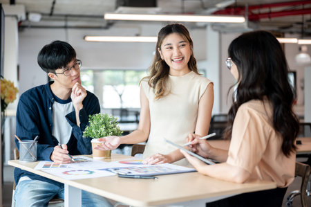 Smiling asian woman designer talking with coworkers about color chart at wooden table in office. Creative business, Corporate job, Modern company.の写真素材