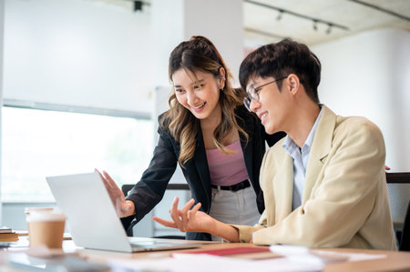 Smiling asian businesswoman looking at laptop talking with intern coworker at wood table in office. Internship Program, Corporate jobs, Modern company.の写真素材