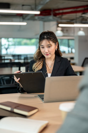 Businesswoman holding clipboard aside laptop sitting across coworker at wooden table in meeting...