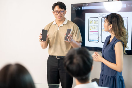 Asian man holding phone talking or presenting app to coworkers in front of monitor at office's...