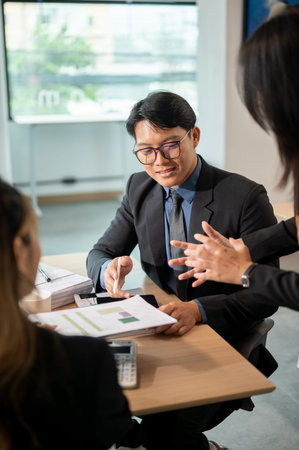 Businessman holding pen reading pointing at document in coworker's hand at wooden table in office....
