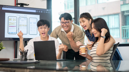 Asian man and coworkers raising fist in excited while looking at laptop on glass table in office....