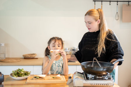 Asian daughter holding and looking at sandwich standing with white mother at kitchen cooking...