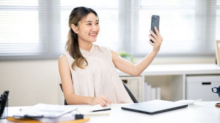 Pretty woman office worker holding smartphone taking a selfie or video calling at table in office....