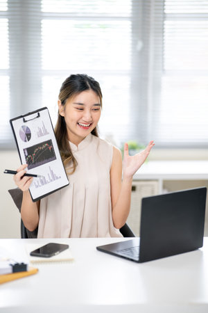 Pretty woman office worker holding pen and clipboard talking in front of a laptop at table in...