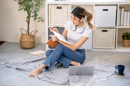 Asian woman holding pen and reading a book aside laptop while sitting on carpet floor in living...