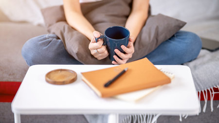 Close up woman's hands holding coffee mug cup aside table with book and coaster while sits on sofa....