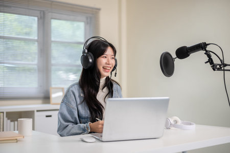 Asian woman hosting a radio show or podcast through laptop with headset and mic at home or office....