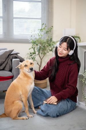 Pretty asian woman with headphones patting puppy dog while sitting on floor carpet in a living room....