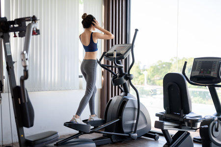 Full body back view shot of a healthy woman wearing headphone while using exercise equipment in gym....
