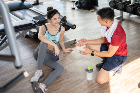 Woman client holding phone talking with fitness trainer as sitting on floor after exercising in gym....