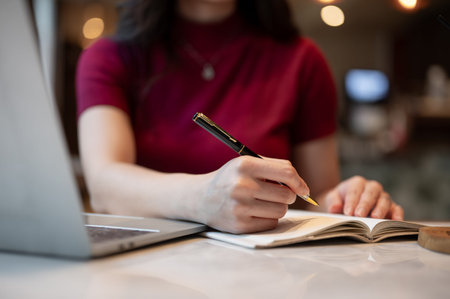 Close up of woman's hand holding pen writing in notebook aside laptop while sitting at table in...