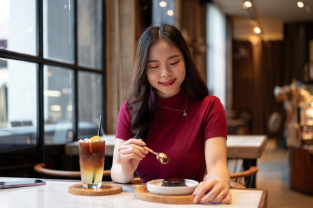 Young asian woman holding spoon eating chocolate cake aside orange coffee cup at table in the cafe....