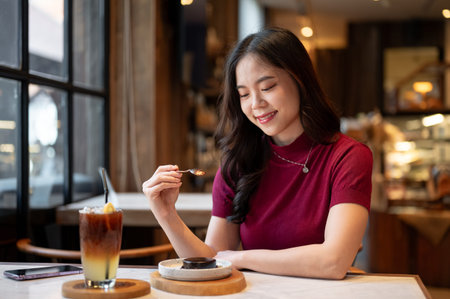 Young asian woman holding spoon eating a chocolate cake aside orange coffee cup at table in cafe....