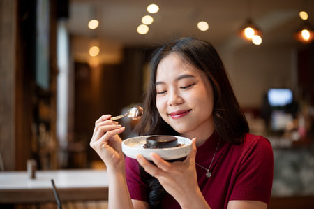 Young asian woman holding silver spoon and smelling a chocolate cake on plate at table in the cafe....