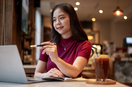 Asian woman holding pen pointing and looking at a laptop aside coffee while sitting at table in...