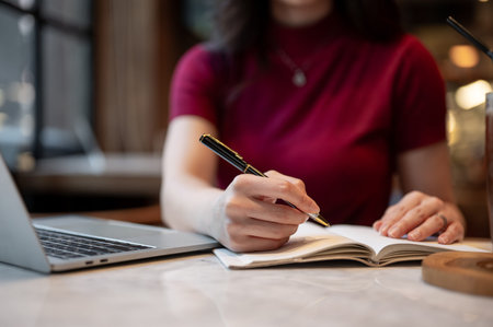 Close up woman's hand holding pen writing in notebook aside laptop while sitting at table in a cafe....