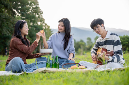 Asian friends woman enjoy eating snacks and man playing guitar sitting on picnic mat in grass field....
