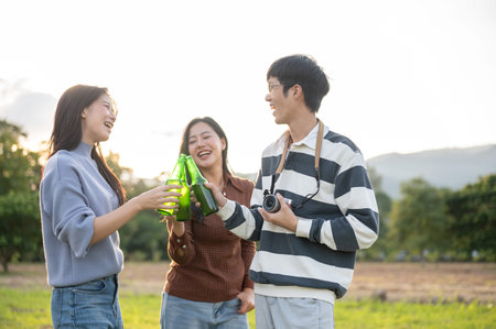 Women and a man friends clinking drink bottles in grass field with mountain view and sunlight. Group...
