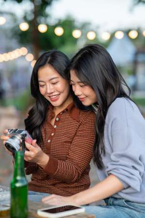 Pretty asian woman holding camera showing pictures to friend at feast table in campground...