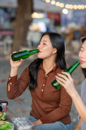 Asian woman and friend drinking alcohol from bottles while sitting at table in campground...