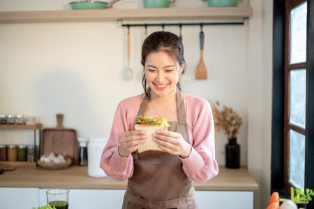 Happy asian woman wearing apron holding and looking proudly at sandwich she makes at kitchen cooking counter. Homemade Food, Self Care, Healthy Living,の写真素材