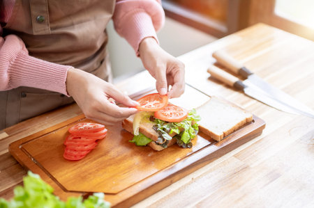 Close up of apron woman putting tomato on bread making veggie sandwich at wooden cooking counter in kitchen. Homemade Food, Self Care, Healthy Living,の写真素材