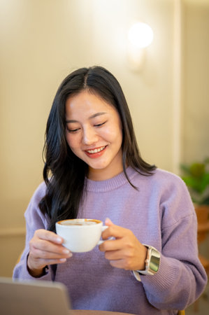 Asian woman holding and looking at latte art coffee cup while sitting across laptop in cafe or shop. Leisure Time, Modern Tech, Chilling Outside.の写真素材