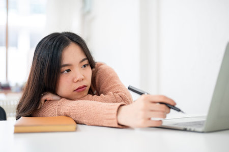 Tired woman resting head on hand holding pen looking at laptop on table in classroom or office....