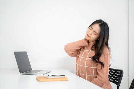 Tired woman massaging her neck in front of a laptop on table in a white classroom or office. Working...