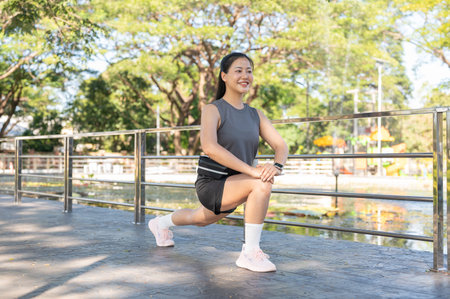 Pretty asian woman in sportswear crouching stretching legs muscles aside railing on pavement in...