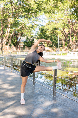 Pretty asian woman in sportswear standing stretching arms and legs over railing on pavement in park....