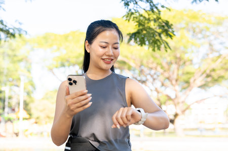Asian woman in sportswear standing holding phone and looking at time on watch while exercise in...