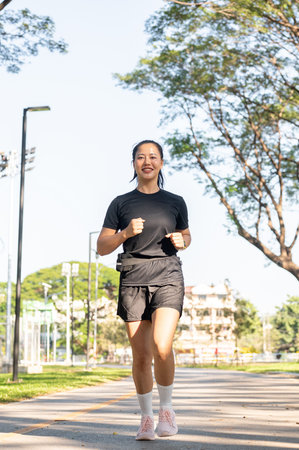 Full body of asian woman in black sportswear exercising running jogging on road in park in sunlight....