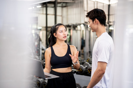Pretty asian woman fitness trainer holding laptop talking to asian man client after exercise in gym....