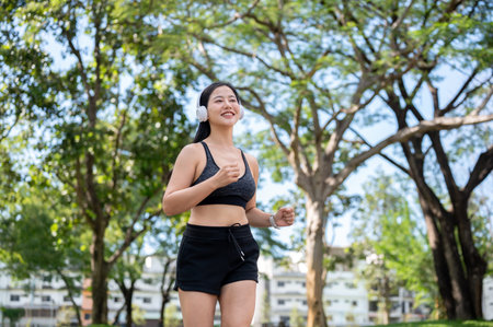 Asian woman in sportswear listening to music in headphones while exercising running jogging in park....