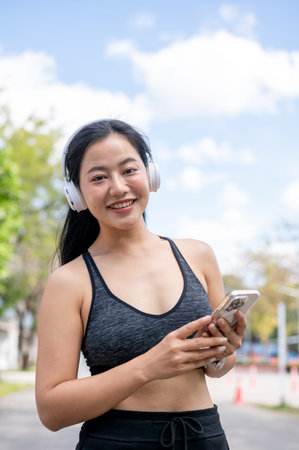 Asian woman in sportswear listening to music in headphones holding a phone while exercising. Healthy living, chilling outdoors.の写真素材
