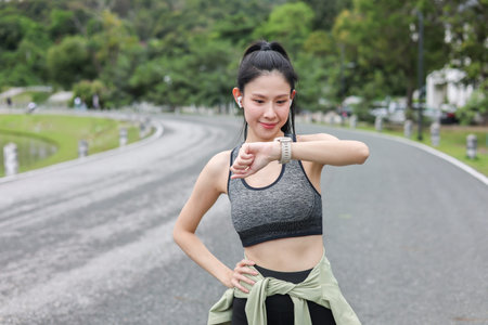 Asian woman in sportswear looking at time on watch before exercise standing on street road in park....