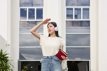Asian woman student holding book using hand covering eye from sunlight standing in front of...