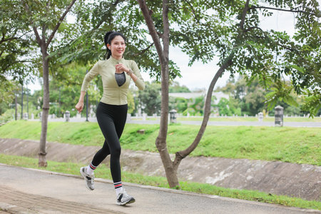 Asian woman in sportswear listening to music while running by the road in park. Healthy Living, Outdoors.の写真素材