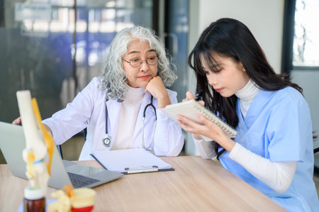 Old asian woman physician with laptop looking at a young doctor taking note sitting at exam table....