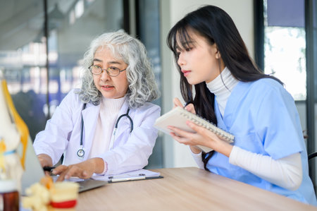 Old asian woman physician typing on laptop showing to young doctor taking note sitting at exam...
