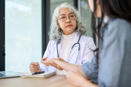 Old asian woman doctor checking pulse on a patient's wrist while sitting at table in diagnosis room....