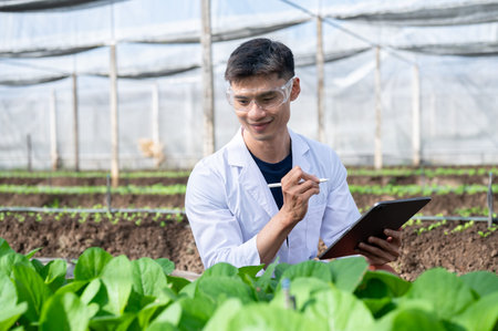 Glasses asian agricultural scientist man holding tablet sitting observing plants in greenhouse farm....