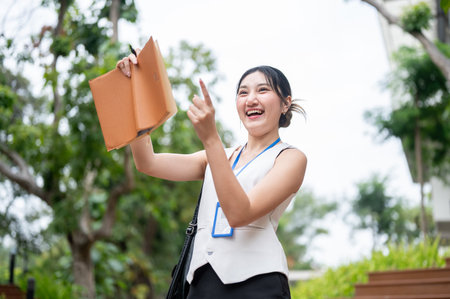 Happy asian woman office worker holding pen and book pointing away while standing or walking in...